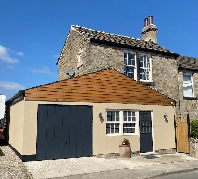 A two-story stone house in Nidderdale features a modern extension with a wooden facade and dark garage door. Nestled in Yorkshire, the building boasts a chimney and a small potted plant by the entrance, offering a charming blend of tradition and contemporary style.